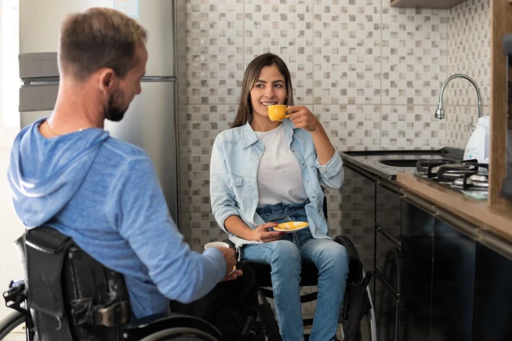 Two disabled friends in wheelchairs at home in kitchen drinking coffee
